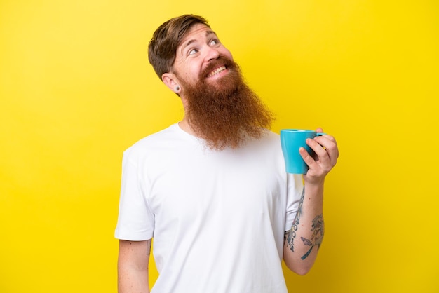 Uomo rosso con la barba che tiene una tazza isolata su sfondo giallo che guarda in alto mentre sorride