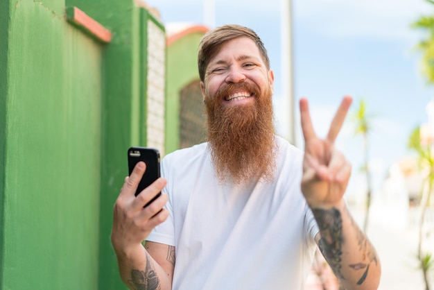 Uomo rosso con la barba che utilizza il telefono cellulare all'aperto sorridendo e mostrando il segno della vittoria