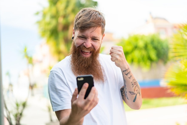 Uomo rosso con la barba che utilizza il telefono cellulare e fa il gesto della vittoria
