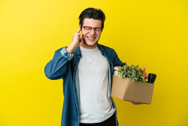 Uomo russo che fa una mossa mentre prende una scatola piena di cose isolate sul muro giallo frustrato e copre le orecchie