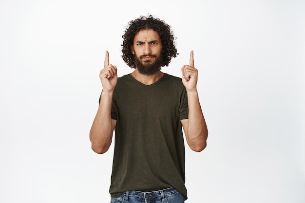 Uomo scettico di Medio Oriente che punta il dito verso l'alto accigliato e guardando deluso esprimere antipatia in piedi in maglietta su sfondo bianco