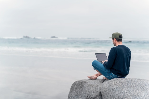 Uomo sconosciuto seduto sulla spiaggia con un computer portatile che lavora vista posteriore