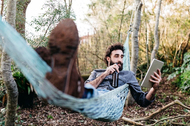 Uomo sdraiato sull'amaca che beve birra e usa il tablet