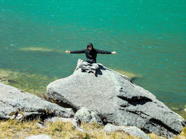 Uomo seduto su una grande roccia nel mezzo di un lago turchese.