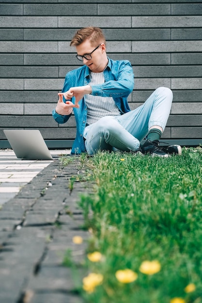 Uomo seduto sull'erba verde e utilizzando il computer portatile sullo sfondo di un muro grigio di casa