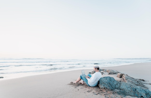 Uomo seduto sulla spiaggia con un laptop da solo che fa telelavoro o lavoro a distanza