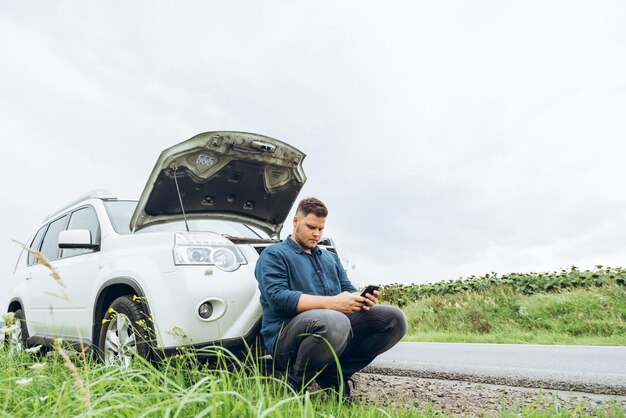 Uomo seduto vicino a un'auto rotta con il telefono, in cerca di aiuto. problemi di viaggio