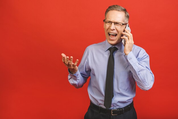 Uomo senior arrabbiato che parla sul telefono cellulare isolato sulla parete rossa. Emozioni negative.