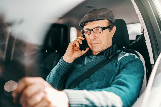 Uomo senior caucasico serio con il cappuccio sulla testa e gli occhiali che si siedono in automobile con la mano sul volante e che per mezzo dello Smart Phone.