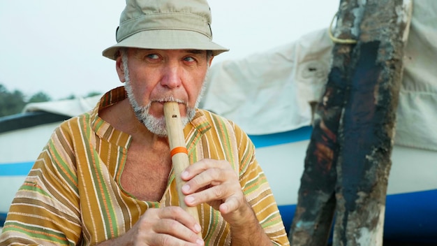 Uomo senior che gioca flauto di bambù sulla spiaggia accanto al peschereccio