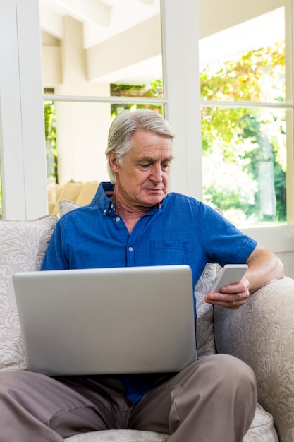 Uomo senior che per mezzo del telefono cellulare mentre lavorando al computer portatile