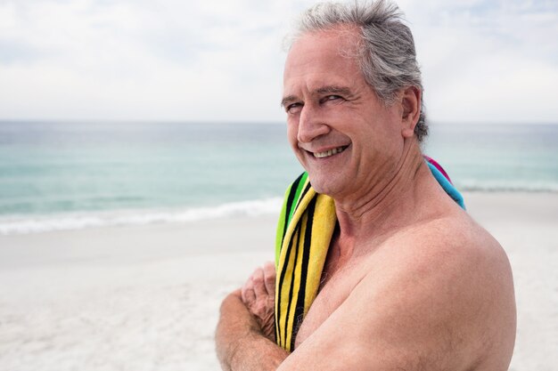 Uomo senior felice che sta sulla spiaggia con l'asciugamano sulla spalla