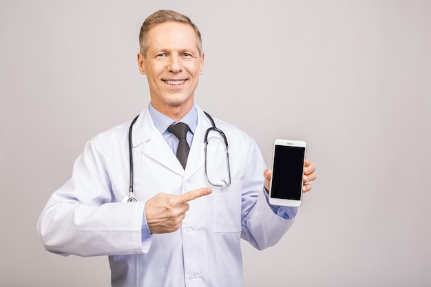 Uomo senior felice di medico che mostra uno schermo in bianco dello Smart Phone isolato su un fondo grigio.