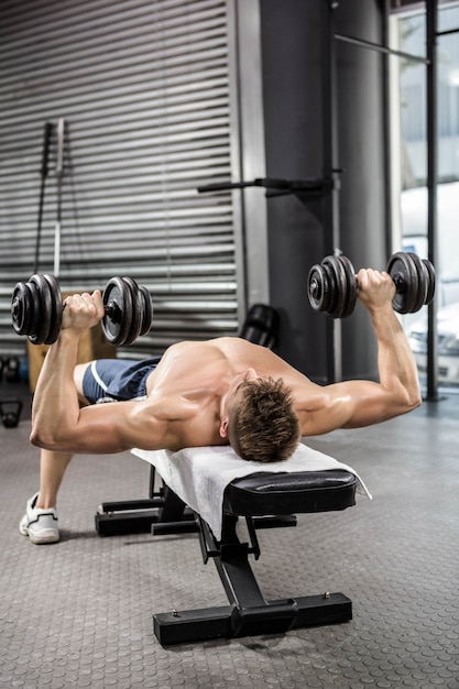 Uomo senza camicia che alza i dumbbells pesanti sul banco in palestra