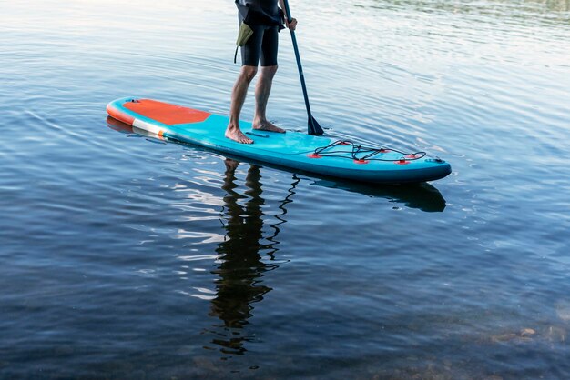 Uomo senza volto in tuta termica che rema in piedi su paddleboard SUP blu sul lago d'acqua con pagaia