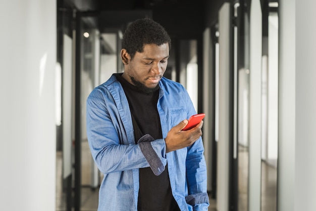 Uomo serio afroamericano che utilizza il telefono cellulare rosso nell'edificio per uffici internet a banda larga g ritratto