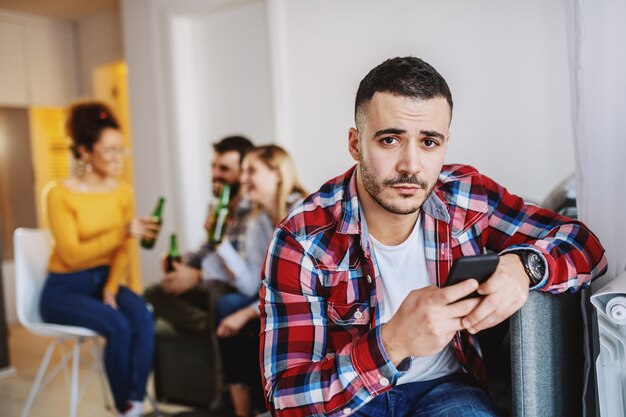 Uomo serio caucasico attraente in camicia a quadri facendo uso dello Smart Phone mentre i suoi amici che chiacchierano e che bevono. Interno soggiorno.