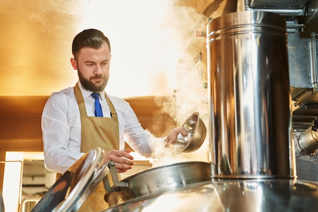 Uomo serio che produce birra. Birraio professionista in camicia bianca e grembiule che lavora nella fabbrica di produzione della birra e controlla il processo di produzione della birra. Concetto di distilleria e bevanda.