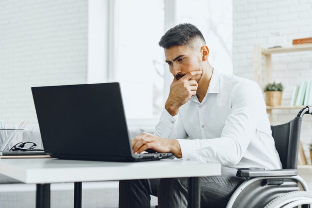 Uomo serio concentrato in sedia a rotelle che utilizza il suo laptop per lavoro / cerca lavoro in Internet