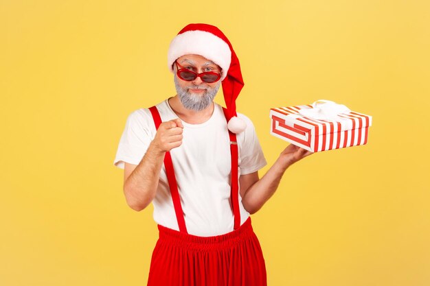 Uomo serio e sicuro di sé in costume e occhiali da sole che tiene la scatola del regalo e punta il dito su di te mentre prepari regali fingendo di essere babbo natale Colpo in studio al coperto isolato su sfondo giallo