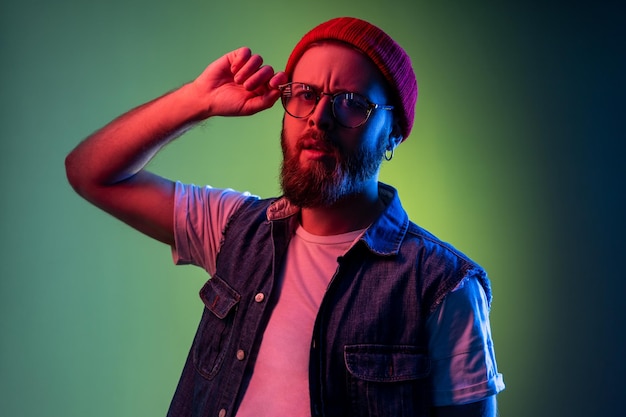 Uomo serio hipster che guarda la telecamera con uno sguardo attento tenendo le braccia sulla montatura degli occhiali che indossa un berretto e gilet di jeans Indoor studio girato isolato su sfondo colorato di luce al neon