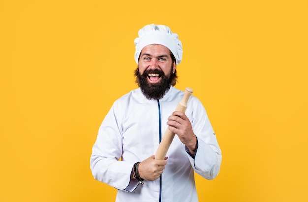 Uomo sicuro dello chef con barba e baffi in uniforme da cuoco che tiene il mattarello su sfondo giallo cucina felice