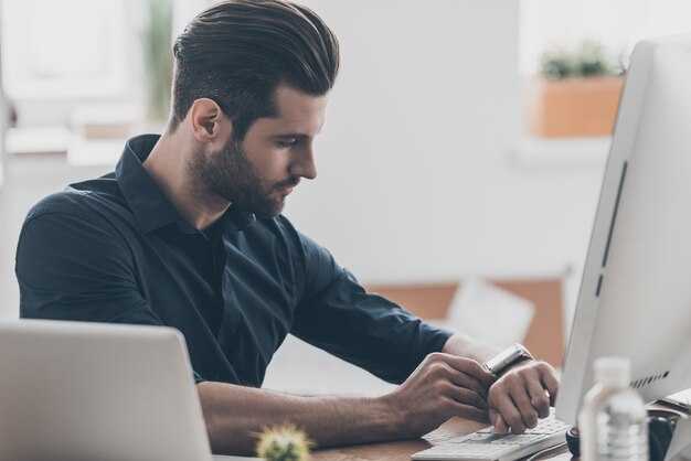 Uomo sicuro di sé. Bel giovane professionista IT che utilizza l'orologio intelligente mentre è seduto alla scrivania in un ufficio creativo