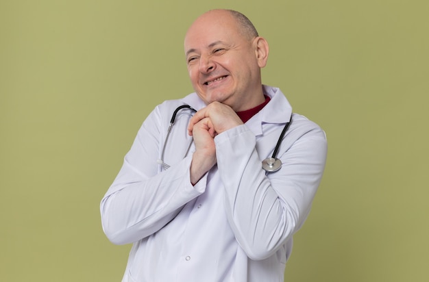 Uomo slavo adulto compiaciuto in uniforme da medico con stetoscopio che si tiene per mano insieme e guarda di lato