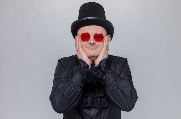Uomo slavo adulto contento con cappello a cilindro e occhiali da sole in camicia gotica nera che si mette le mani sul viso