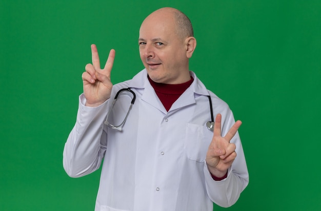 Uomo slavo adulto impressionato in uniforme da medico con lo stetoscopio che gesturing il segno di vittoria