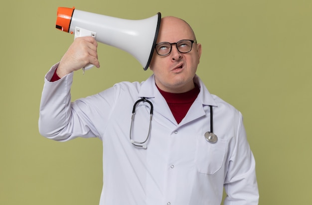 Uomo slavo adulto infastidito con occhiali ottici in uniforme da medico con stetoscopio che tiene altoparlante e rotea gli occhi