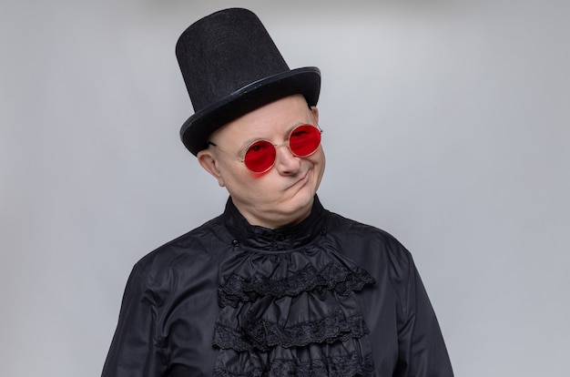 Uomo slavo adulto premuroso con cappello a cilindro e occhiali da sole in camicia gotica nera che guarda in alto