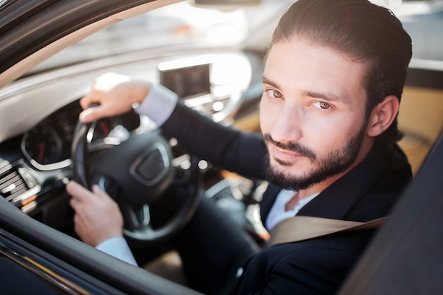 Uomo soddisfatto e felice che si siede in automobile e posa. Guarda la telecamera e sorride un po '. Il ragazzo barbuto tiene le mani sul volante.