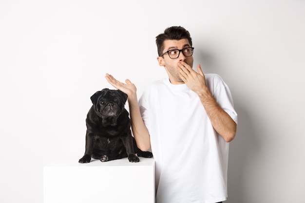 Uomo sorpreso e scioccato con gli occhiali, in piedi vicino a un simpatico cane nero e fissando il logo, in posa su sfondo bianco.