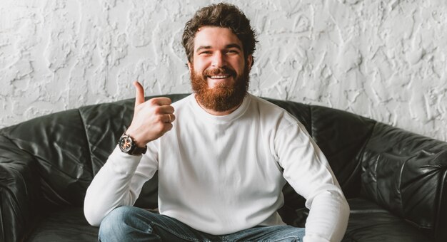 Uomo sorridente allegro con la barba che si siede sul sofà e che mostra i pollici su