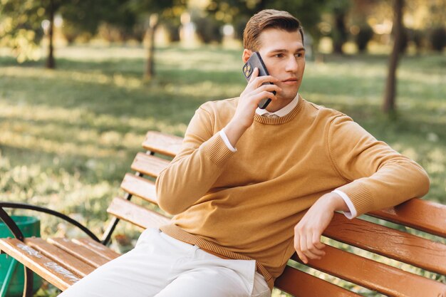 Uomo sorridente bello moderno in abiti gialli e bianchi che parla al telefono seduto su una panchina del parco al tramonto