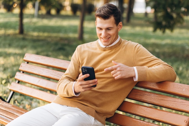 Uomo sorridente bello moderno in abiti gialli e bianchi che parla al telefono seduto su una panchina del parco al tramonto