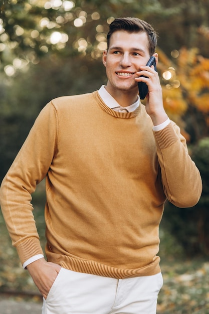 Uomo sorridente bello moderno in vestiti gialli e bianchi che parla al telefono nel parco al tramonto