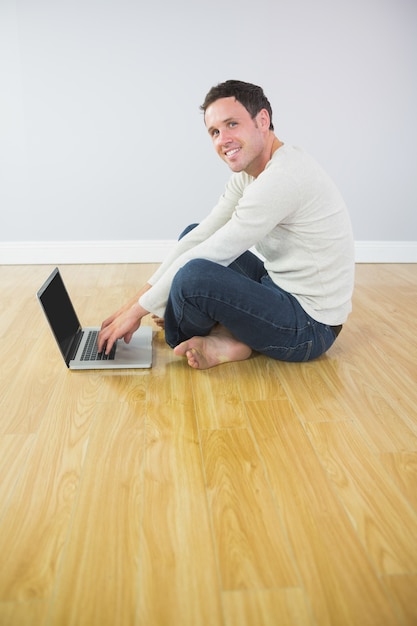 Uomo sorridente casuale che si siede sul pavimento per mezzo del computer portatile