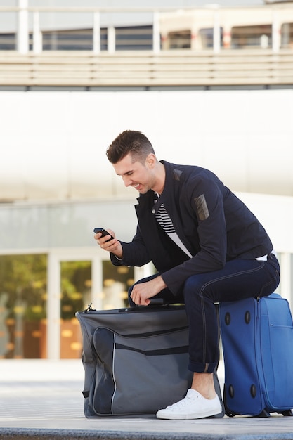 Uomo sorridente che aspetta con i bagagli e telefono alla stazione