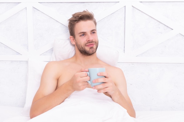 Uomo sorridente che beve il tè dalla tazza in camera da letto bianca sul letto