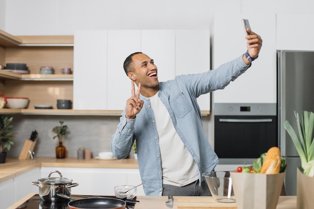 Uomo sorridente che fa selfie o fa una videochiamata mentre cucina cibo vegano sano all'interno della cucina
