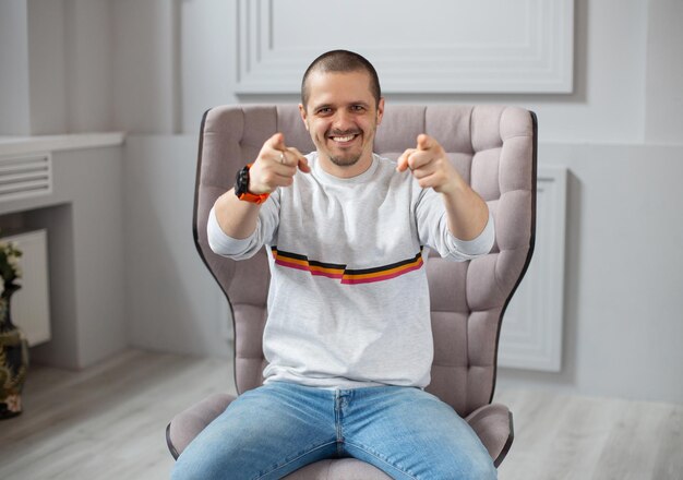 Uomo sorridente che indica con due mani in camera mentre è seduto su una sedia o un divano in una stanza moderna