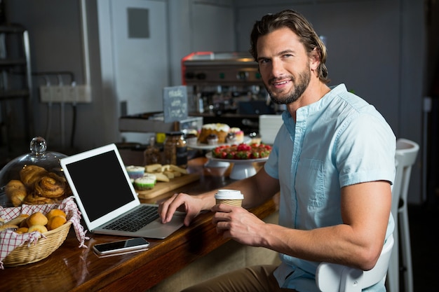 Uomo sorridente che mangia caffè mentre si utilizza il computer portatile al bancone