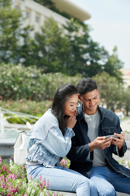 Uomo sorridente che mostra video divertenti sullo smartphone alla sua ragazza quando sono seduti nel parco