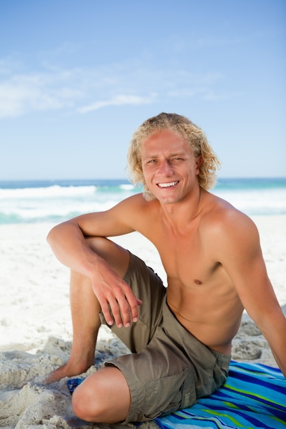 Uomo sorridente che si siede sul suo asciugamano da spiaggia mentre guardando la telecamera