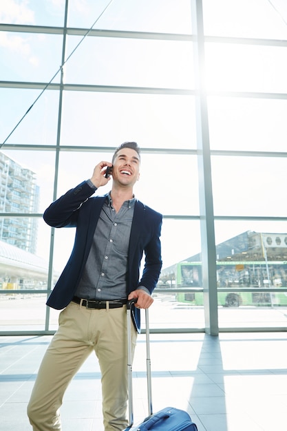 Uomo sorridente che sta nella stazione con il telefono cellulare e la valigia