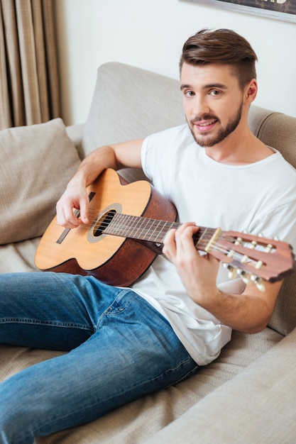 Uomo sorridente che suona la chitarra sul divano e guarda davanti