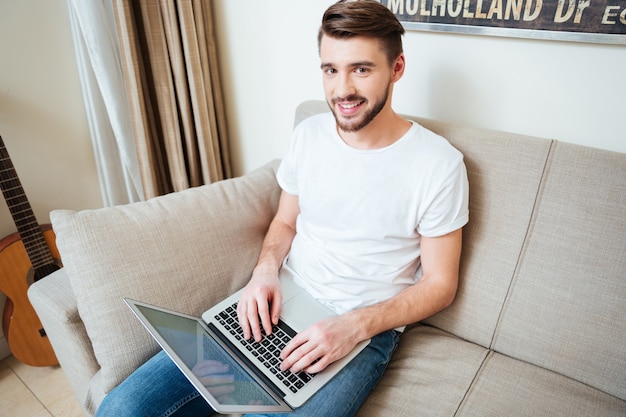 Uomo sorridente che usa il computer portatile sul divano di casa e guarda davanti