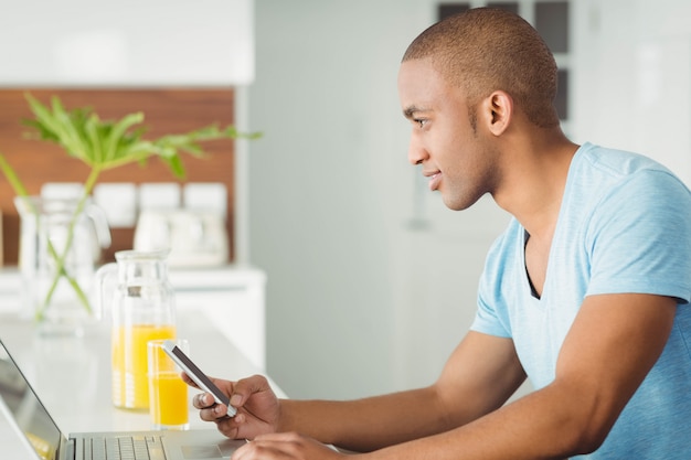 Uomo sorridente che utilizza computer portatile e smartphone nel salone a casa